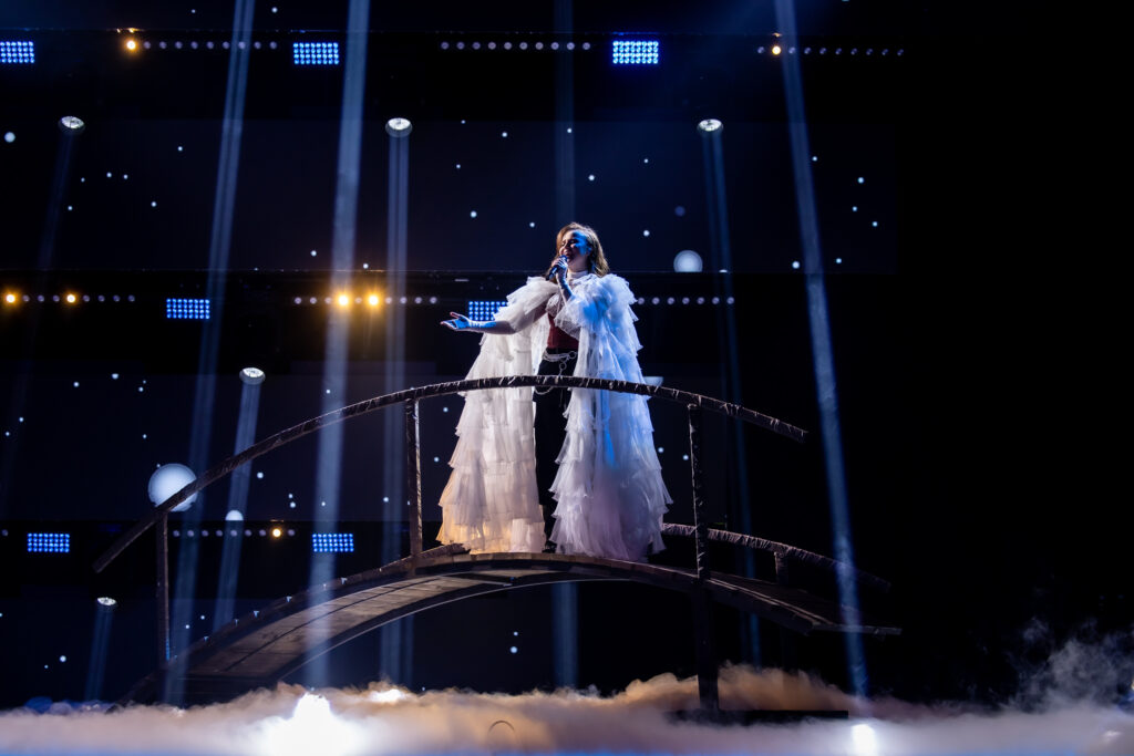 Kuvassa valkoiseen höyhenasuun pukeutunut tummahiuksinen nainen. Laulaa lavalla olevan siltarakennelman päällä mikrofoniin.